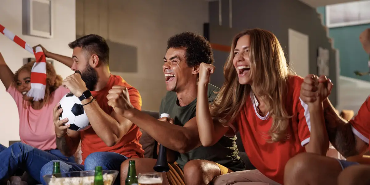 Group of people sat on a sofa watching sports. Our of shot would appear to be a television. The group are excited about the action unfolding on the television.