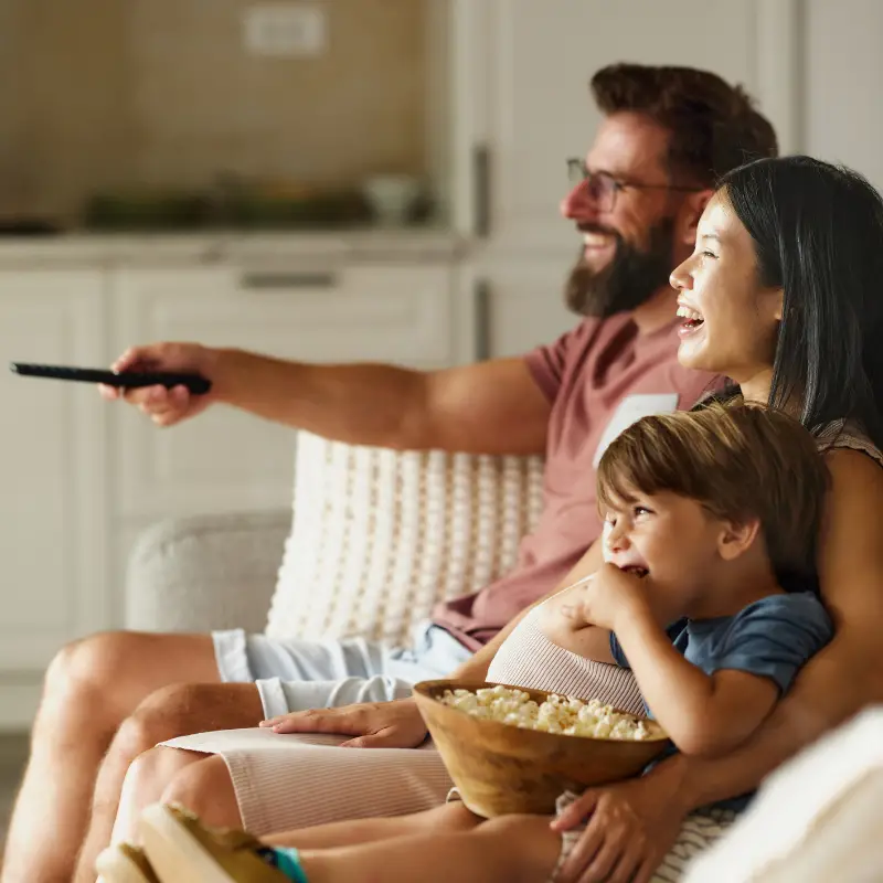 A family on a sofa. An adult male has a remote control in his hand, indicating there is a television out of shot. A young child is holding a bowl of popcorn.